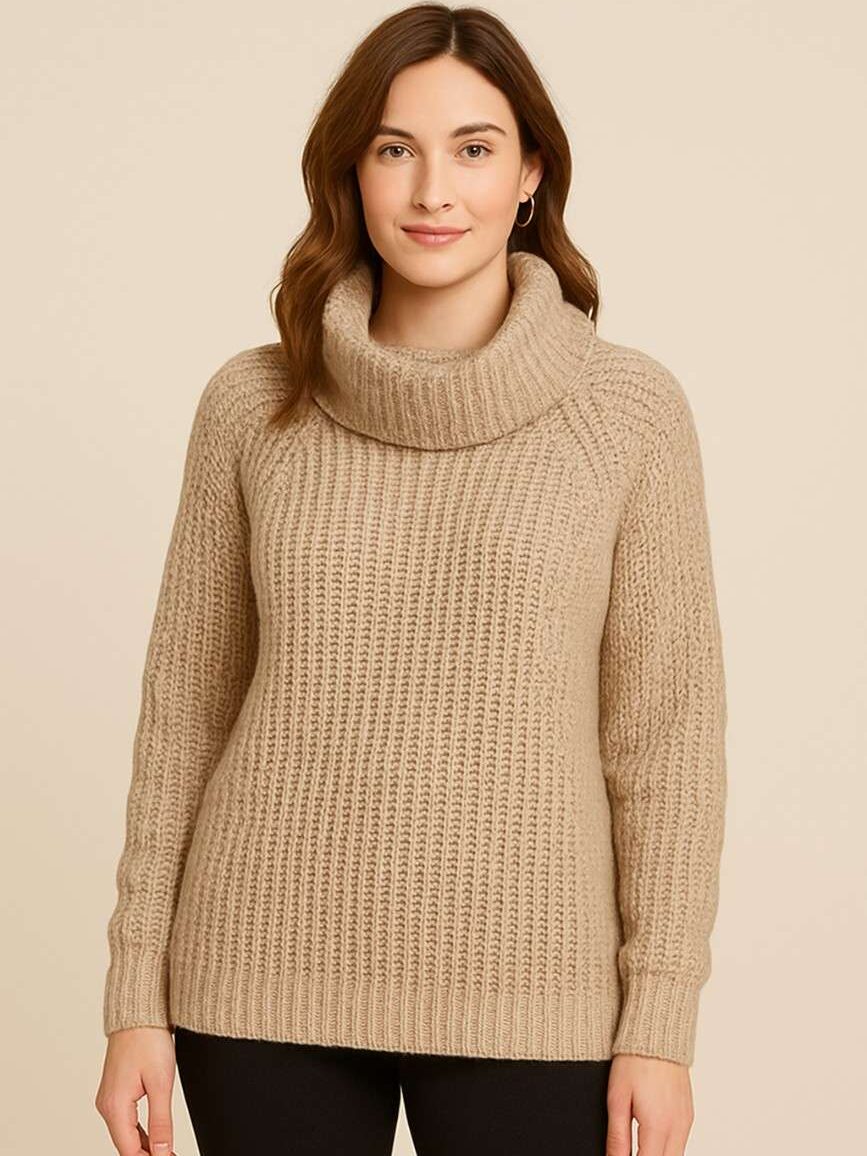 A young woman with fair skin wears a cozy beige chunky knit cowl neck sweater paired with black leggings. She stands confidently against a soft beige background, exuding warmth and relaxed winter style.